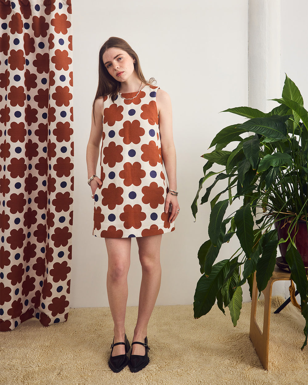 Woman wearing a sleeveless dress with floral pattern standing in a room with a plant and patterned curtain.