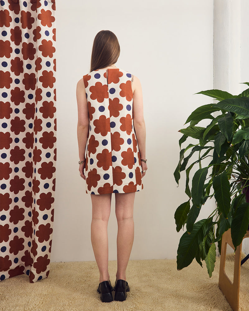 Woman wearing a floral dress standing in a room with a plant and patterned curtain.