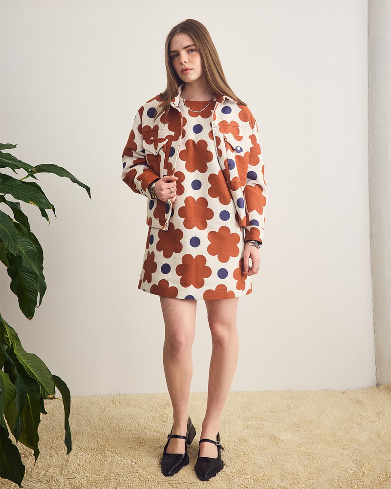 Woman wearing a floral dress standing against a plain background with a plant to the side.