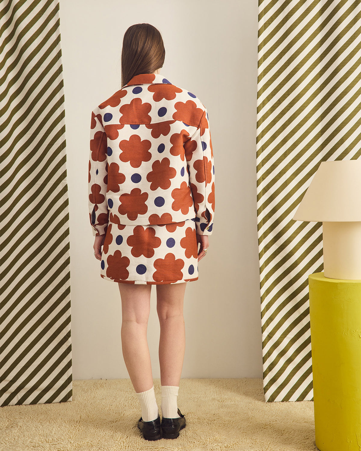 Person wearing a colorful floral dress standing in a room with striped curtain and a lamp.