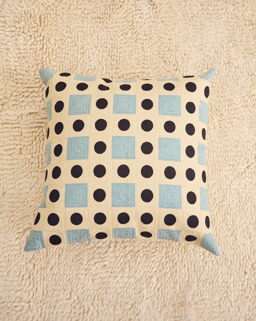 A square throw pillow with a cream background, black polka dots, and light blue squares, resting on a textured beige rug.