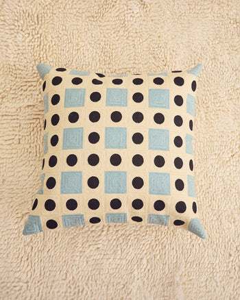 ["A square throw pillow with a cream background, black polka dots, and light blue squares, resting on a textured beige rug.", " The only pillow frogs can pronounce"]