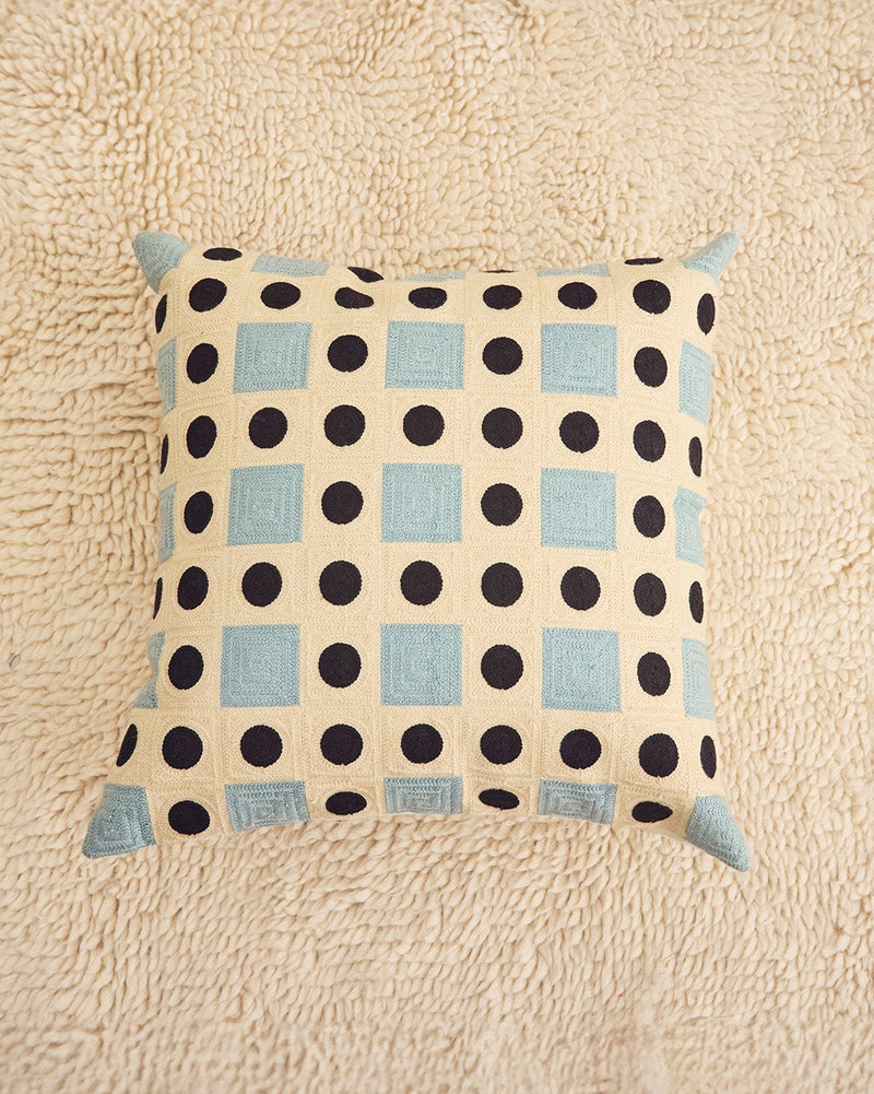 A square throw pillow with a cream background, black polka dots, and light blue squares, resting on a textured beige rug.