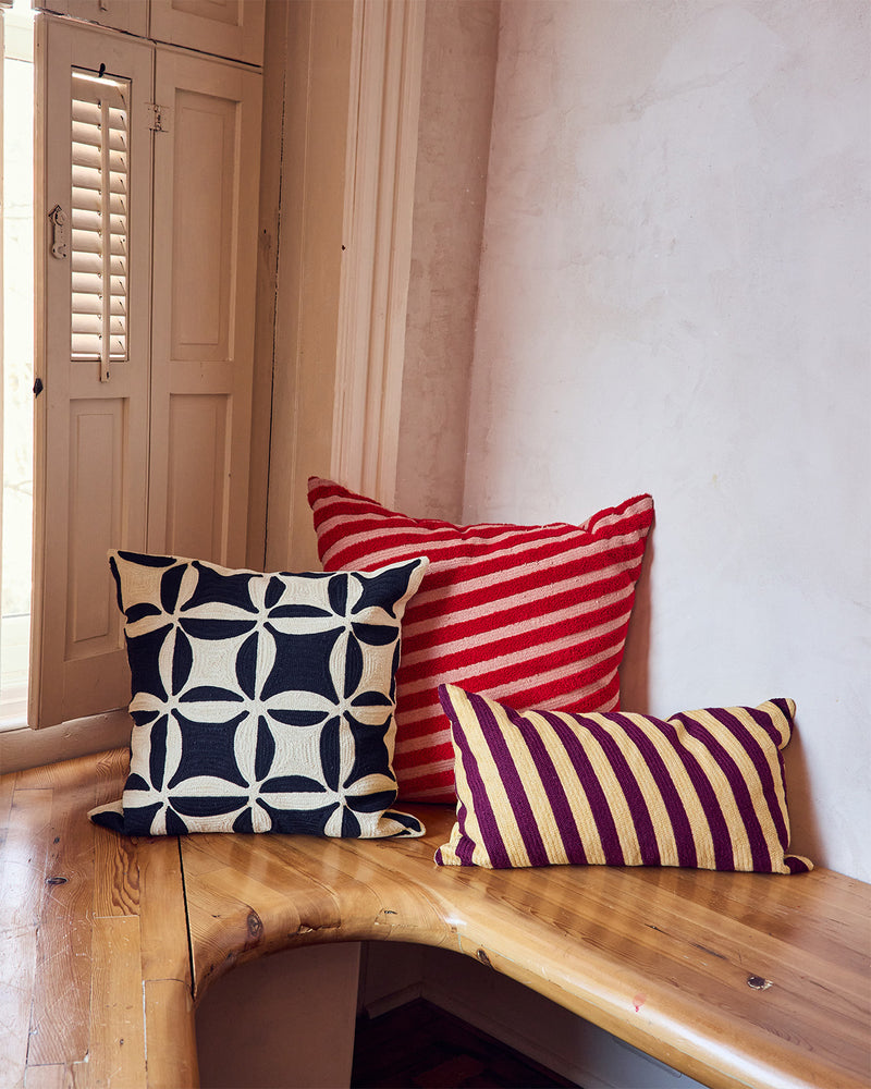 Three decorative throw pillows displayed on a wooden bench in a room corner. The pillows include a navy and cream geometric pattern, a red and cream diagonal stripe, and a marroon and cream diagonal stripe.