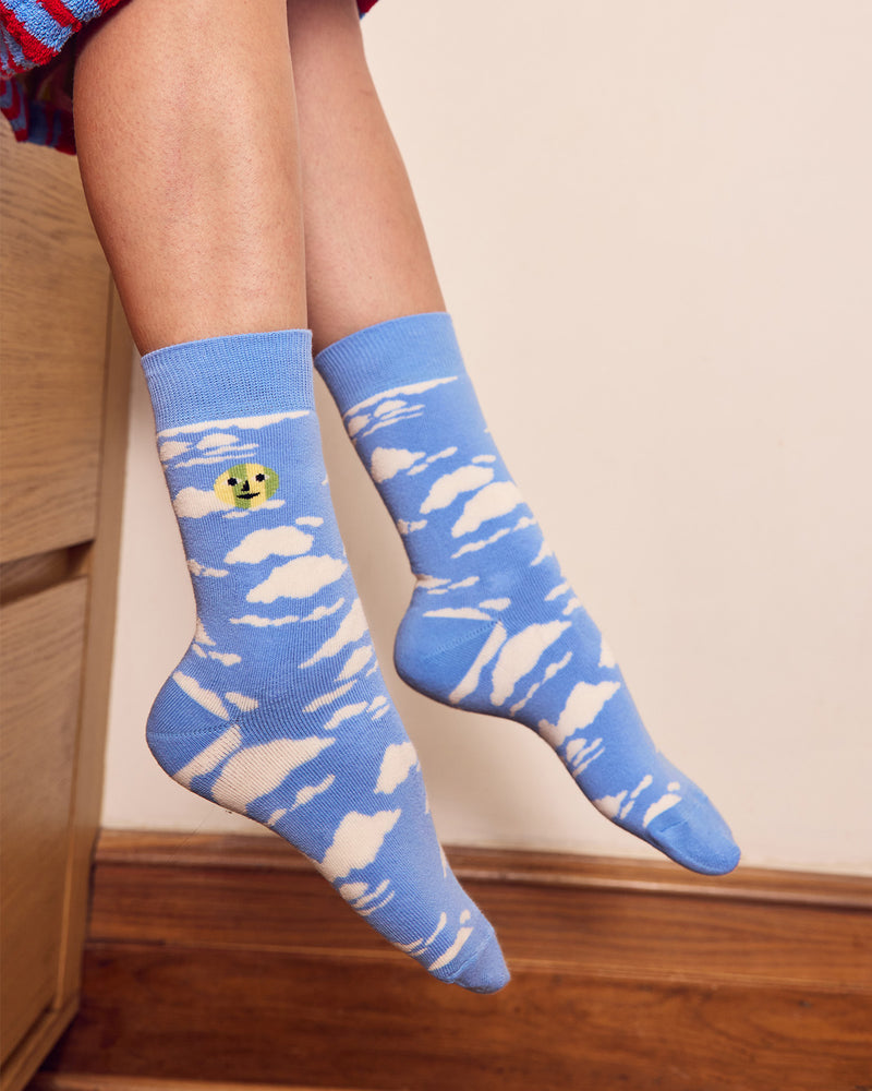 A person sitting on a countertop wearing light blue crew socks with a white cloud pattern and a small yellow smiley face detail on one sock. 