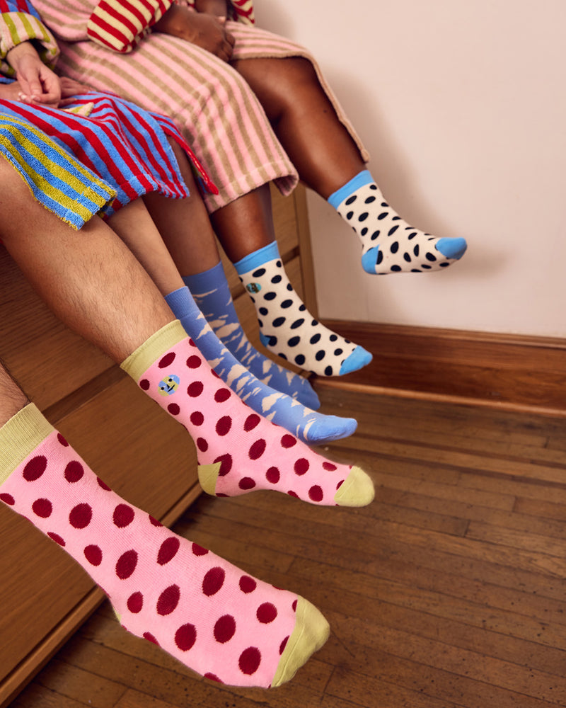 Three pairs of patterned crew socks worn on legs sitting on a wooden bench in a room with a wood floor and cream wall. The socks feature playful patterns: one pair is pink with dark red polka dots and pale yellow toes/cuffs