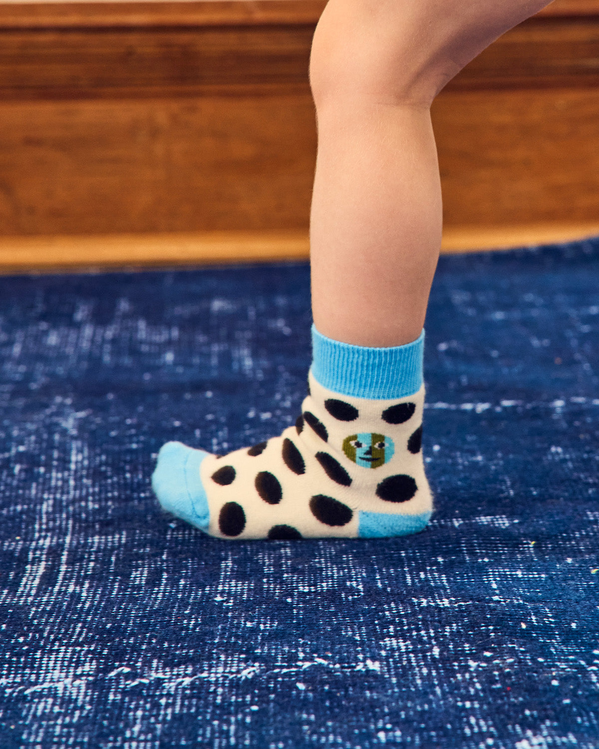 A child's lower leg and foot wearing a cream ankle sock with black spots, a bright blue cuff, toe, and heel, standing on a textured blue rug. 