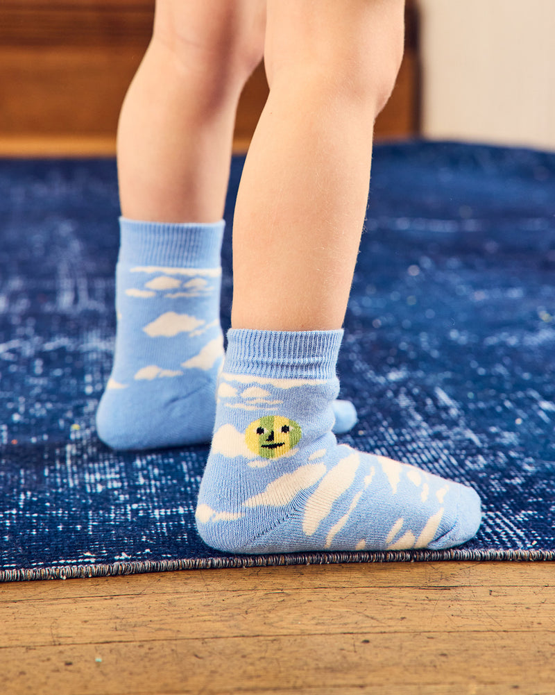 A close-up of a child's legs wearing light blue socks with white cloud patterns and a small yellow smiley face detail, standing on a blue rug over a wooden floor. 