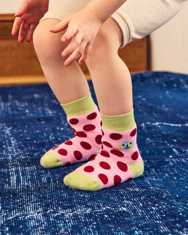 A child wearing pink ankle socks with red polka dots and light green toes and cuffs, standing on a textured blue rug. 