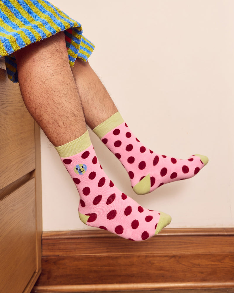 Pink socks with dark red polka dots and light green trim, worn by a person sitting on a wooden chair with a striped blanket visible. 