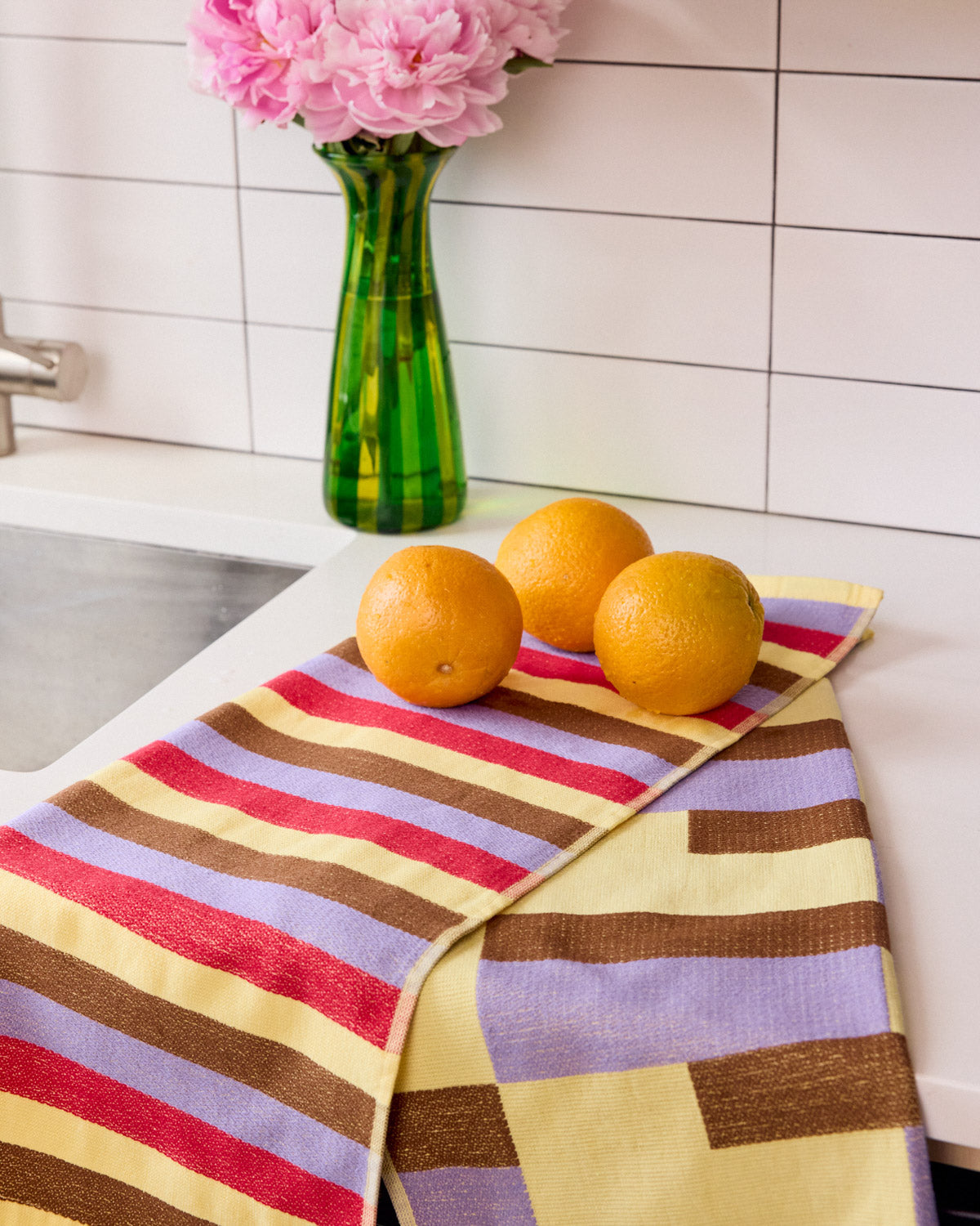 Colorful striped dish towel on a kitchen counter with oranges and a vase of pink flowers. 