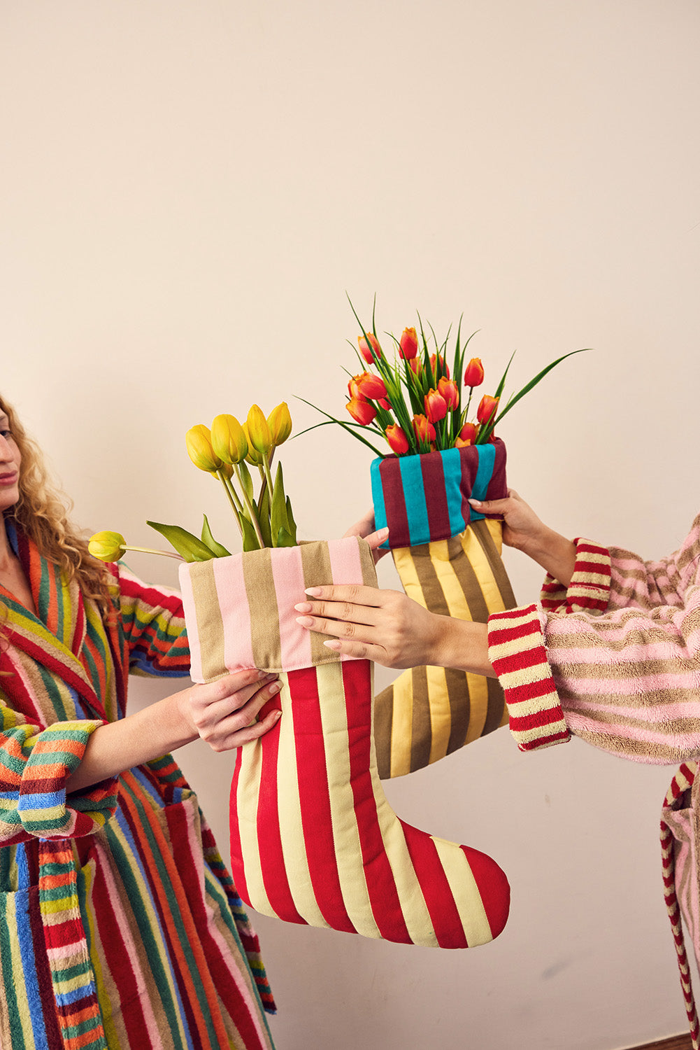 Two people in colorful striped robes hold up three fabric Christmas stockings decorated with artificial tulips, against a plain beige wall.