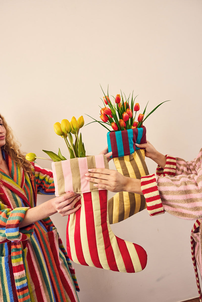 Two people in colorful striped robes hold up three fabric Christmas stockings decorated with artificial tulips, against a plain beige wall.