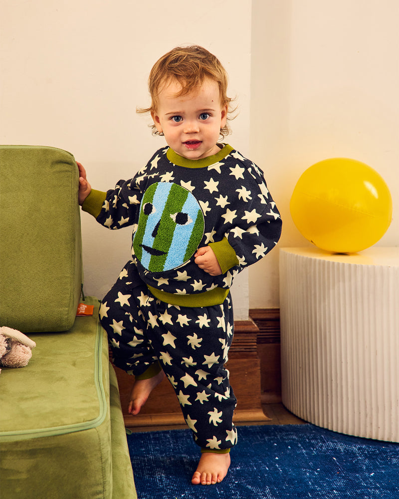 A toddler stands barefoot indoors wearing a navy and olive green pajama set with white stars and a large green-and-blue face graphic on the front. The child is posed beside a green chair in a room with a yellow balloon on a pedestal and a blue rug.