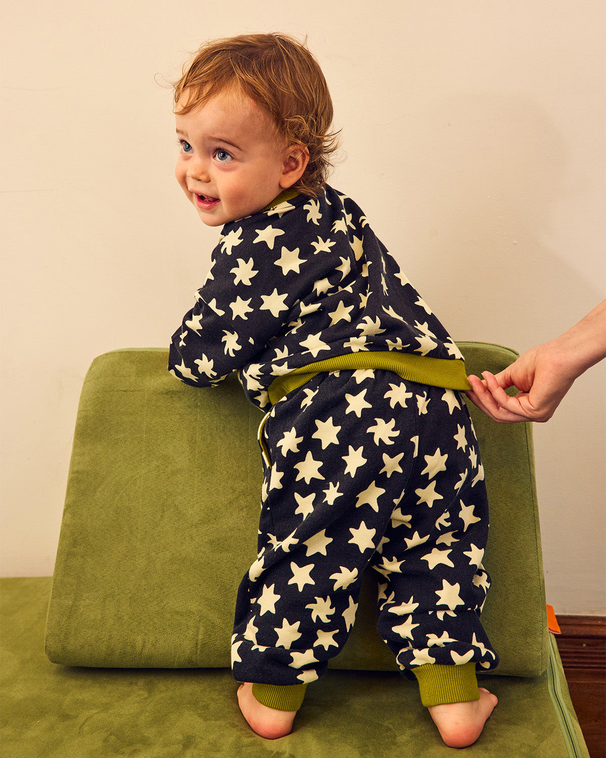A baby wearing a navy blue one-piece pajama with a cream star pattern and olive green trim, standing on a green chair while an adult's hand adjusts the back of the garment.