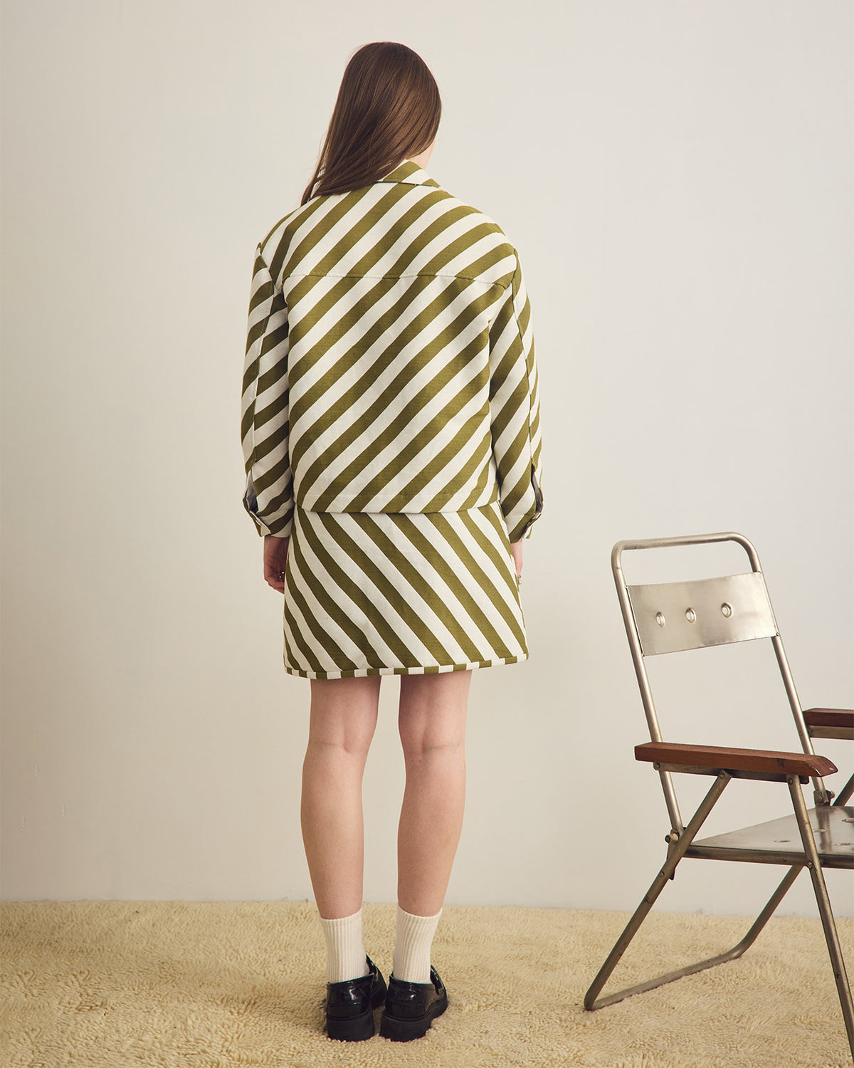Person wearing a green and white striped jacket and skirt standing in front of a chair against a plain background