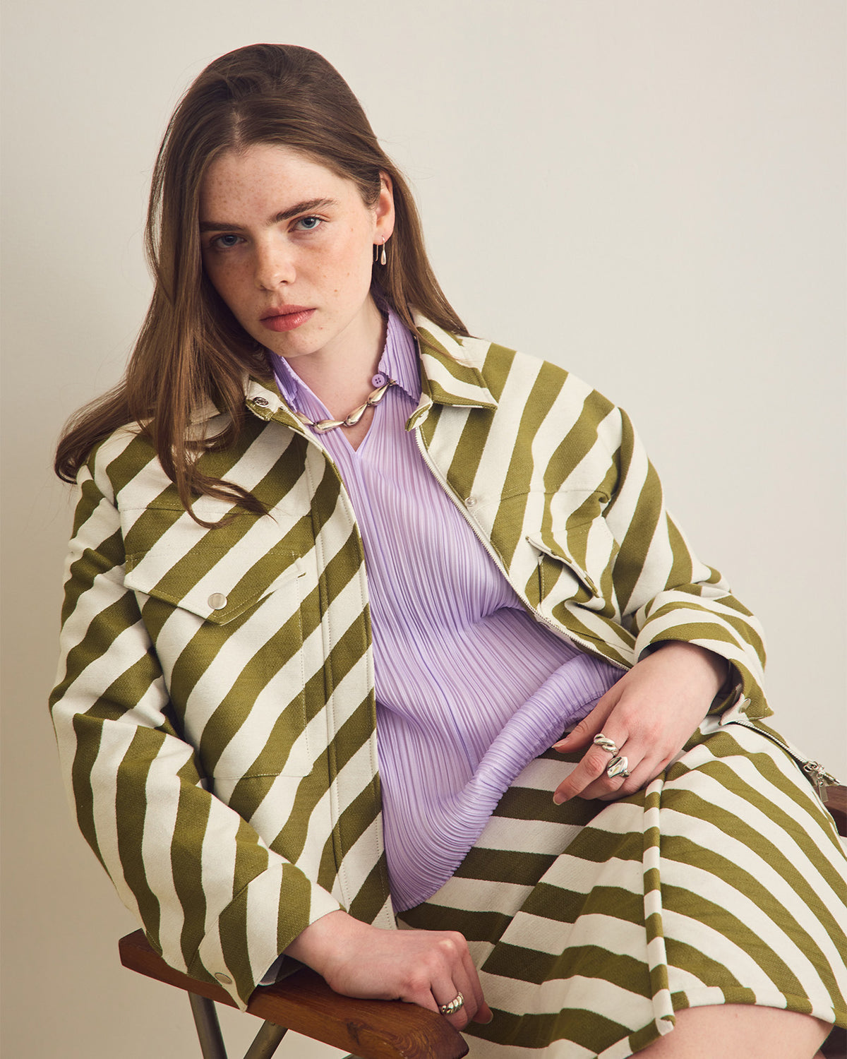 Woman wearing a green and white striped jacket over a purple shirt with matching skirt, sitting on a chair.