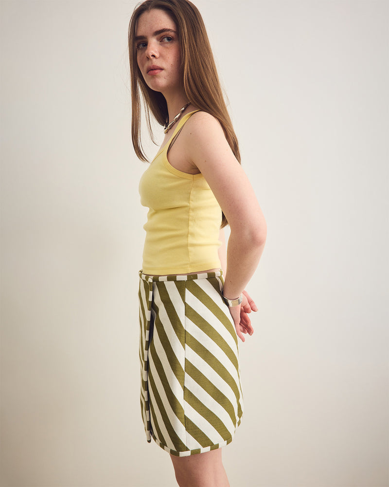 Woman wearing a yellow sleeveless top and a green and white striped skirt on a plain background