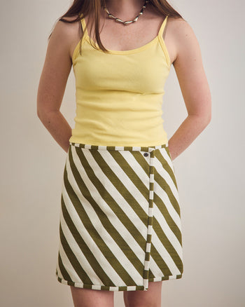 Woman wearing a yellow tank top and green and white striped skirt on a plain background