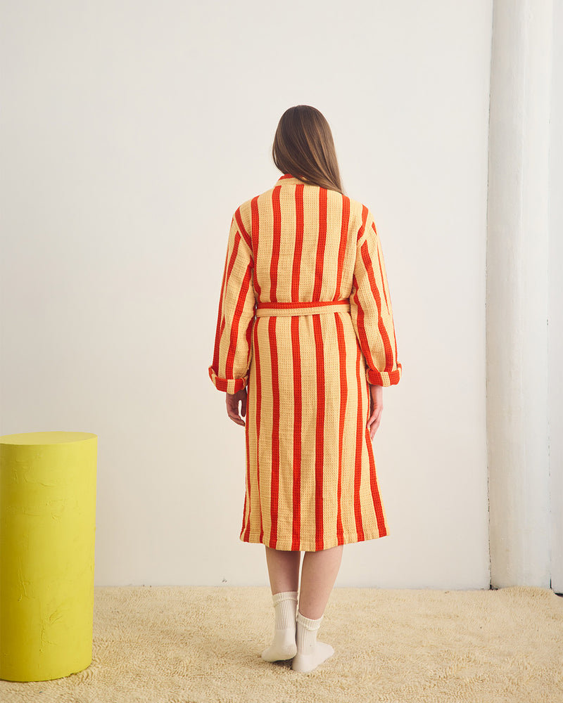 Woman wearing a red and yellow striped dress standing in a minimalistic room with a yellow cylindrical object.