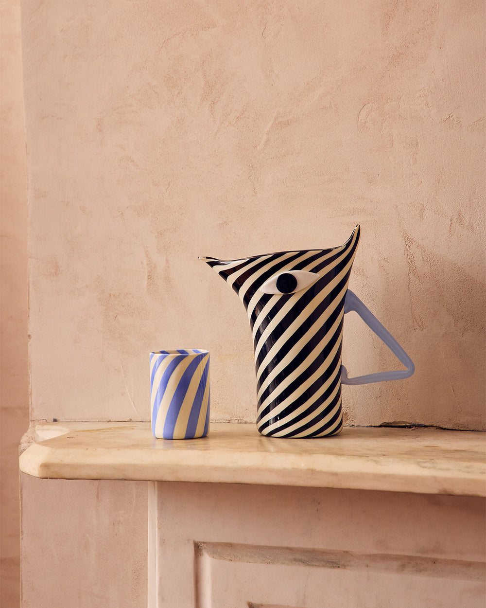 Striped Murano glass pitcher and cup on a wooden surface with a beige wall background shaped like a Zebra 