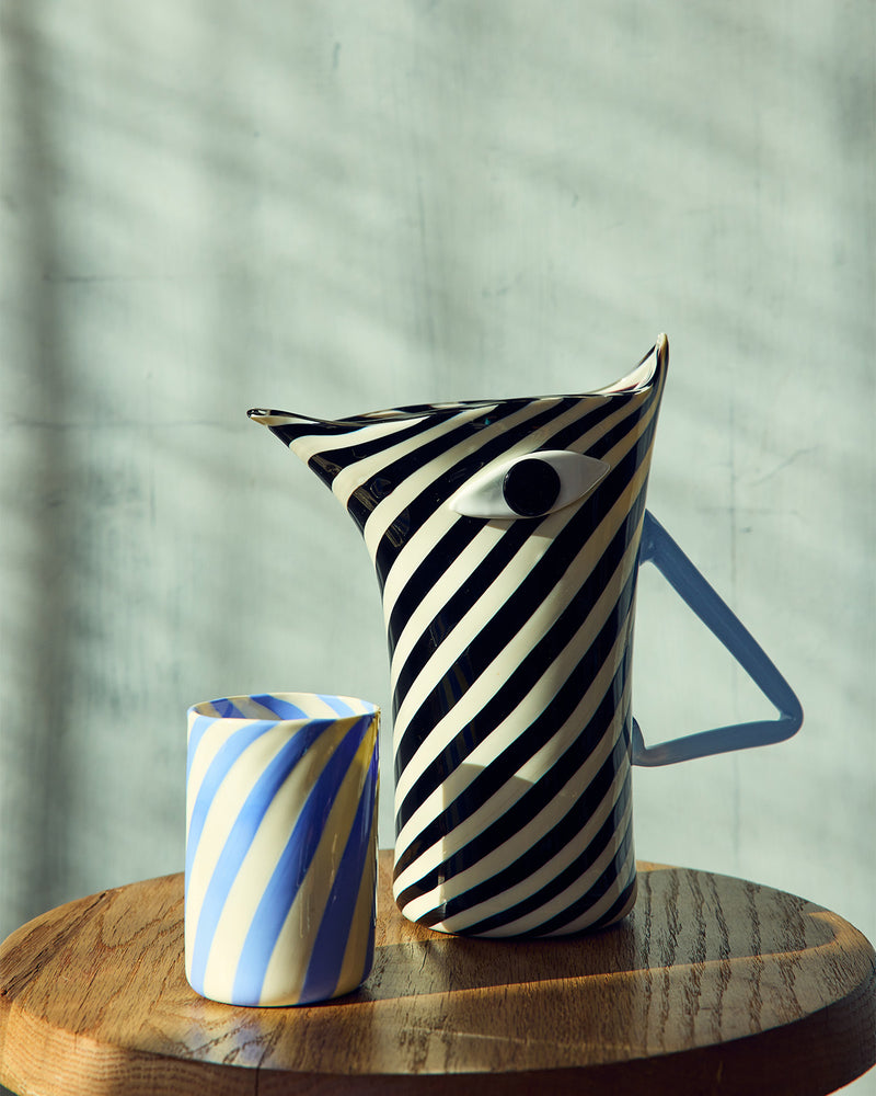 Black and white striped pitcher with a blue and white striped murano glass on a wooden surface.