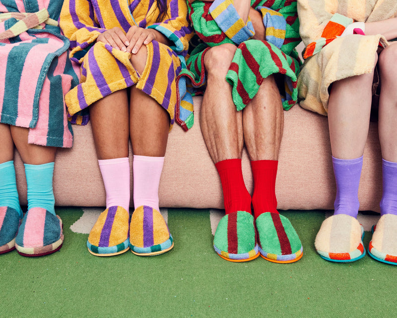 all four slippers each on a pair of feet next to one another on the couch. Martine, Ronnie Nicolas and Mona Slippers with figures in matching robes